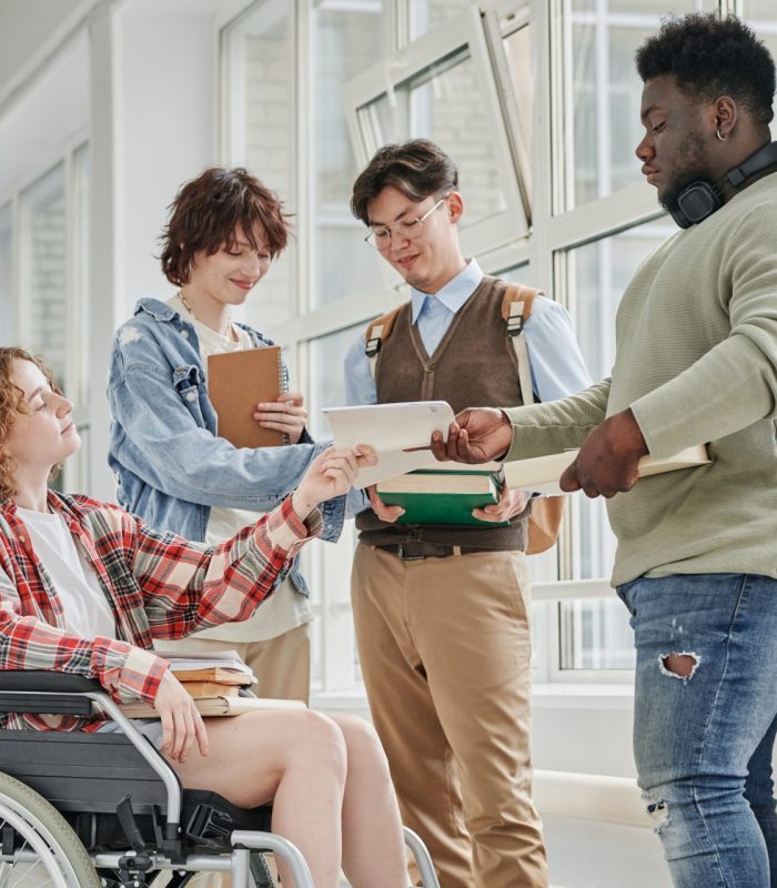 Cute teenage girl with disability taking copybook of African American guy standing in front of her while their classmates having discussion