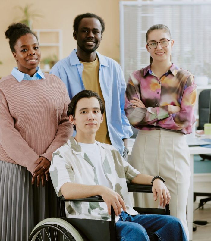Young IT specialist in wheelchair surrounded by his young biracial coworkers while posing for portrait in comfort office