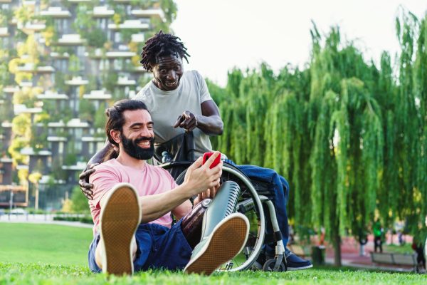 Joyful friends, one with a prosthetic leg and the other in a wheelchair, sharing a laugh in a sunny park setting.