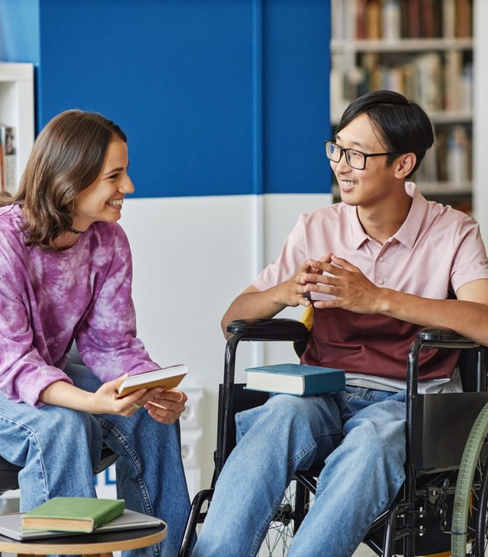 Portrait of smiling young man in wheelchair talking to friend while studying together in library