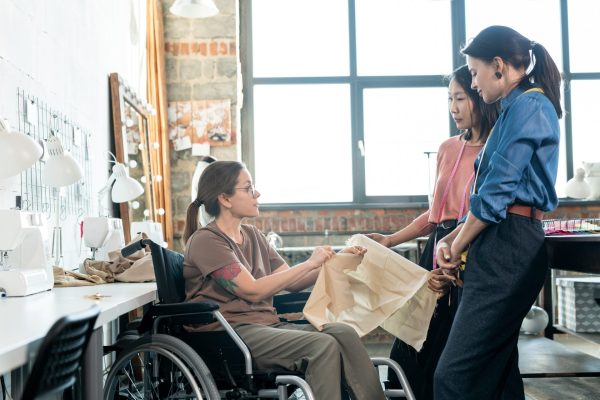 Young fashion designers talking to seamstress in wheelchair in workshop